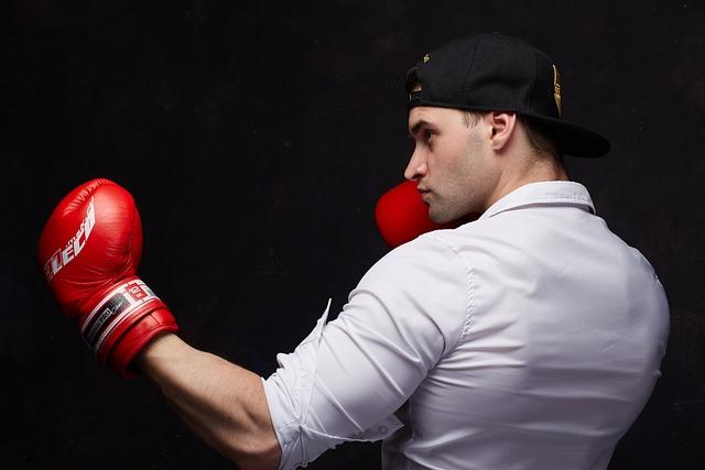 A young man with a backward baseball cap and a button up shirt and boxing glove standing in a boxing stance.