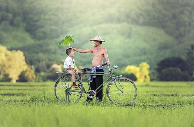 An Asian man holding a bike and touching the top of the head of his son who is sitting on the back of the bike.