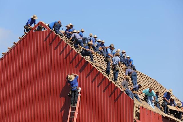 About 25 men on the roof of a building they are building or fixing. One man is at the top of a ladder, everyone else is sitting or standing on the roof itself.