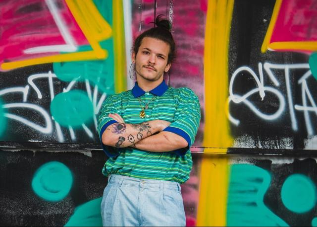 A young man with a top-bun and folded arms in front of a wall of grafitti.