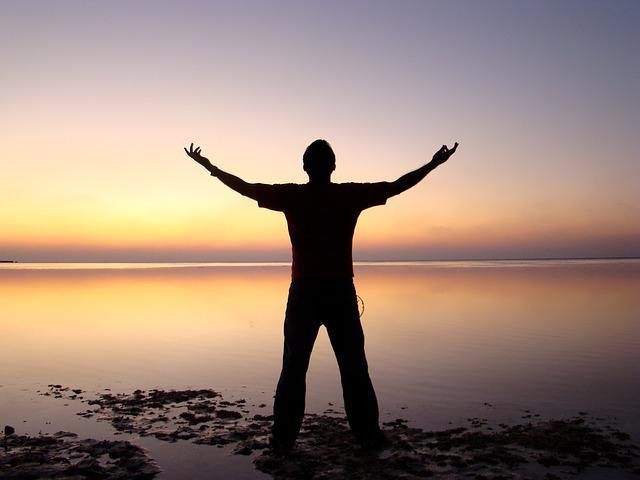 silhouette of a man standing by a lake with his hands stretched up to the sky