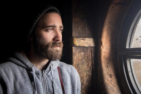 Man in early 30s looking out a window with a pensive expression.