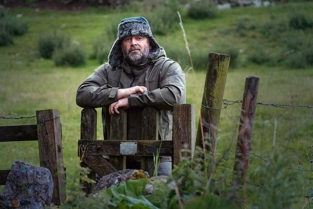 Man wearing a hat with ear flaps standing at an old wooden gate that is slightly ajar.