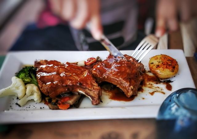 Ribs and vegetables on a rectangular white plate being eaten with a knife and fork.