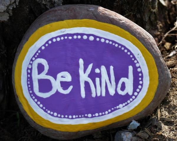 The words Be Kind on a round rock painted in purple and white with a yellow frame. 