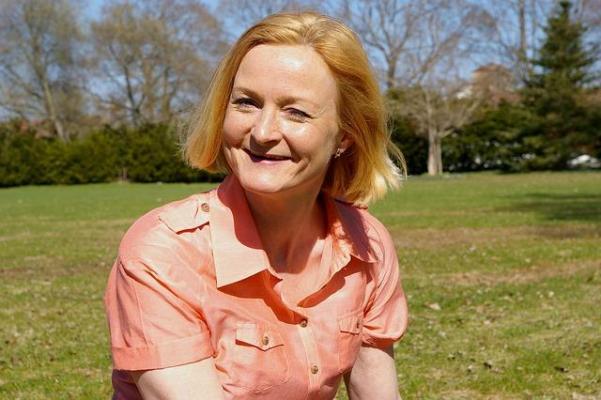 Middle-aged woman with short blonde hair sitting in a grassy field looking directly at us with a smile.