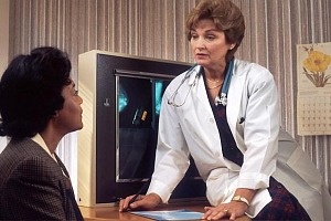 A female doctor sitting on a desk speaking with another woman sitting in a chair.