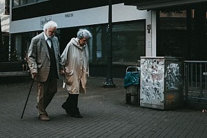 An old couple walking along a sidewalk holding hands, heads down, he is using a cane.