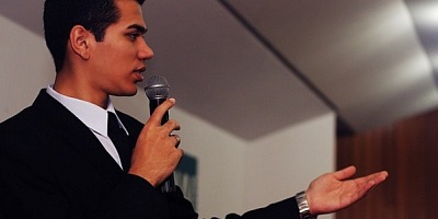 A young dark-haired man with a mic in his hands, preaching to a congregation.