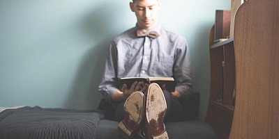 A young man sitting on a bed, leaning against the wall, legs crossed, shoes on, reading a book.